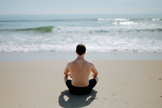 Man meditating on a sandy beach facing the ocean.